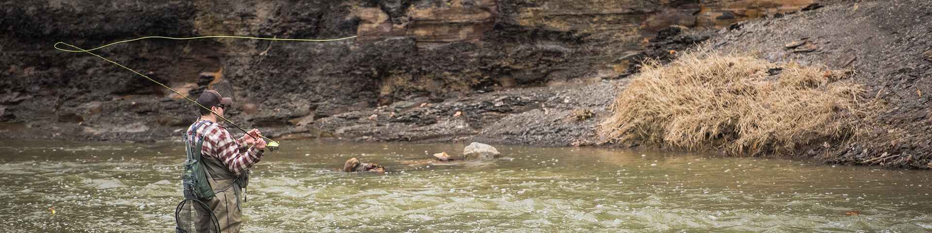 An angler dressed in waders and a plaid shirt is fly-fishing in a rocky river, casting a line with precision. The riverbank is steep with layered rock formations and patches of dry vegetation.