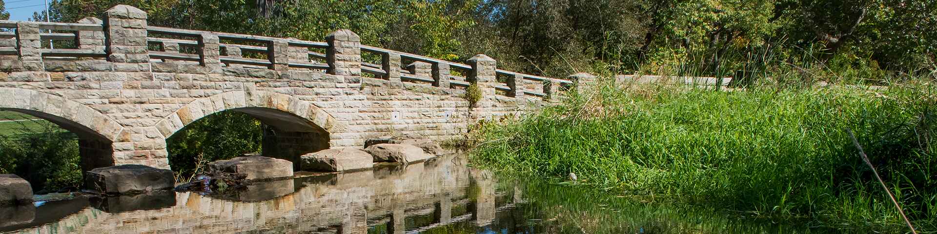 A stone bridge crosses over a calm stream, surrounded by green vegetation and trees under a clear blue sky.