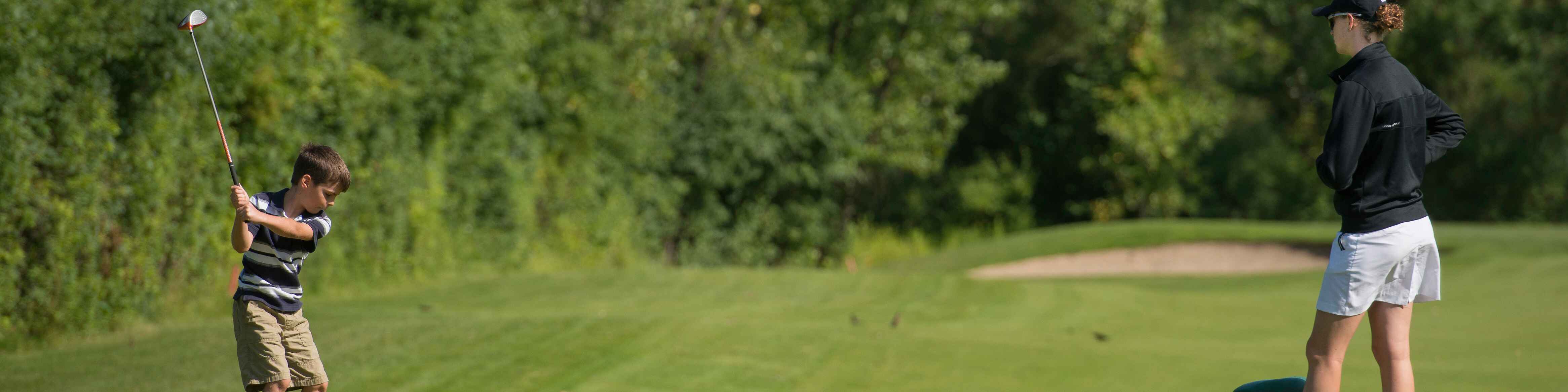 A young boy taking a swing with a golf club, with a coach standing nearby.