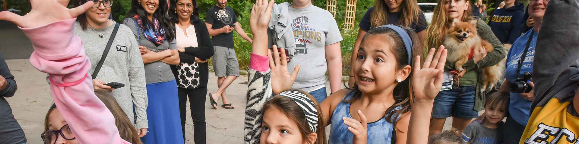 A group of smiling children and adults gathered outdoors, with some kids enthusiastically raising their hands and a small dog being held by one of the adults in the background.
