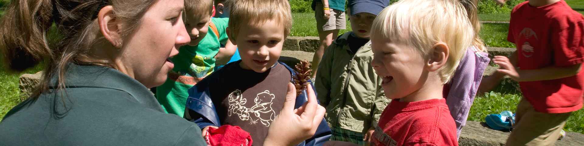 A group of young children outdoors, observing a pine cone held by a guide.
