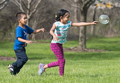Two children delighting in bubble play in a vibrant park, with lush greenery and a clear blue sky in the background.