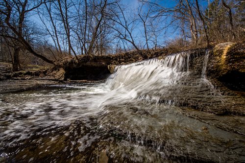 arafed waterfall flowing over rocks in a wooded area, a tilt shift photo by Mike Bierek, featured on cg society, environmental art, water cascading, cascading waterfalls, wide angle landscape photography