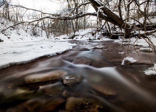 A picturesque stream meanders through a snowy forest, flanked by rocks and trees, embodying a peaceful winter scene.
